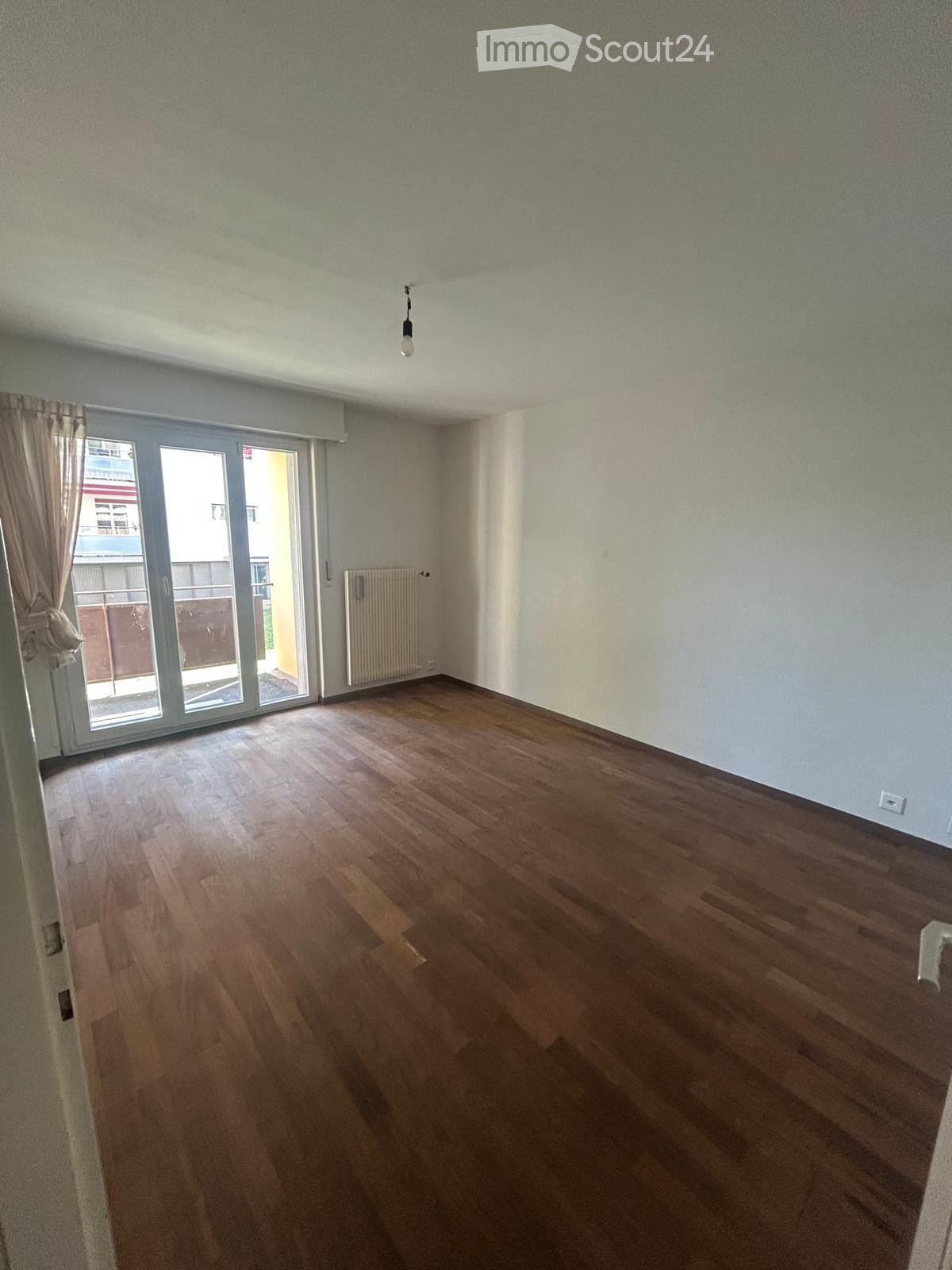 Empty room with wooden flooring, large sliding glass door leading to a balcony, radiator, and a single light fixture on the ceiling.