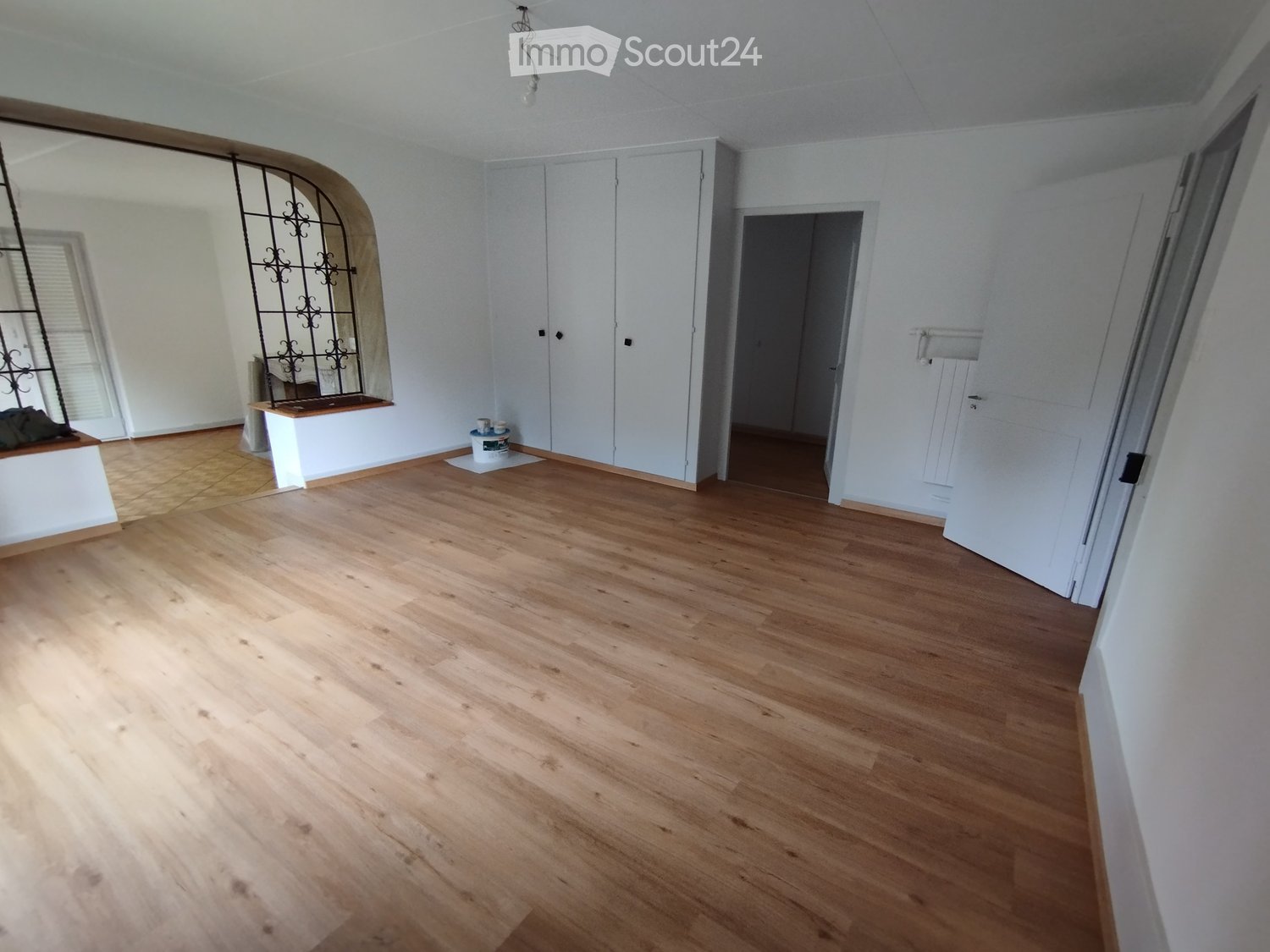 Empty room with light wooden flooring, white walls, and a large arched window with decorative metal bars. A small bench and a few construction materials are visible near the window.