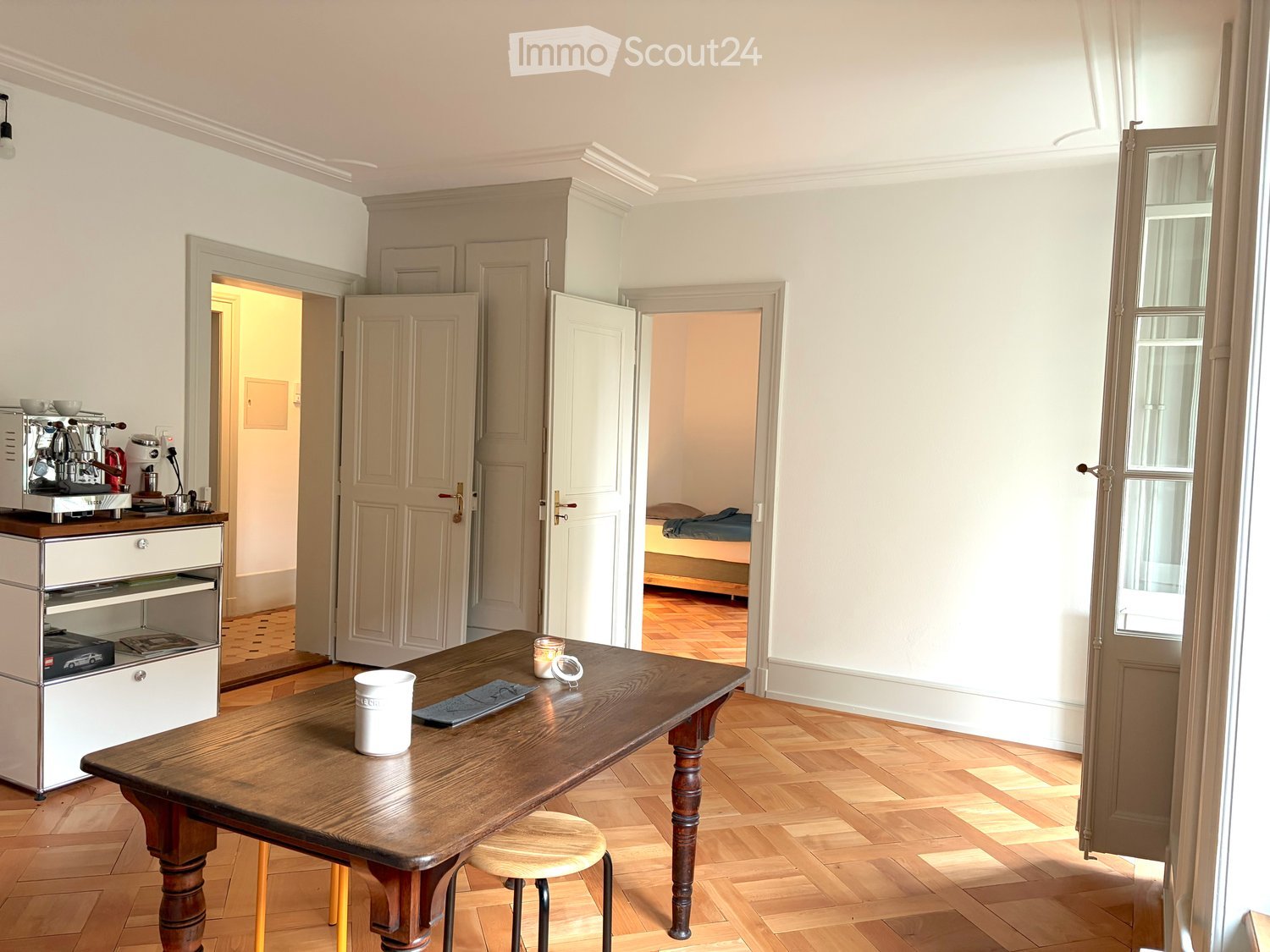 A well-lit kitchen area with a wooden table, a stool, a coffee machine, and an oven. The room features light-colored walls, wooden flooring, and several open doors leading to other rooms.