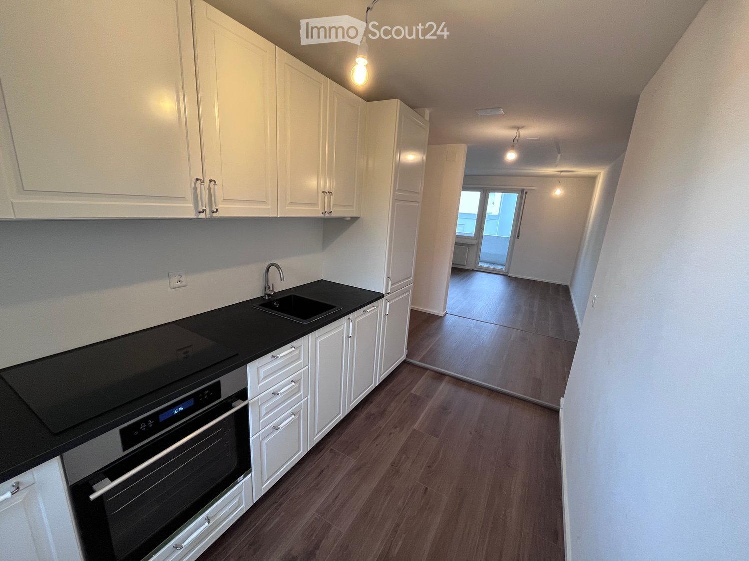 Modern kitchen with white cabinets, black countertop, sink, and built-in oven. The floor is wooden, and the space is well-lit with pendant lights.