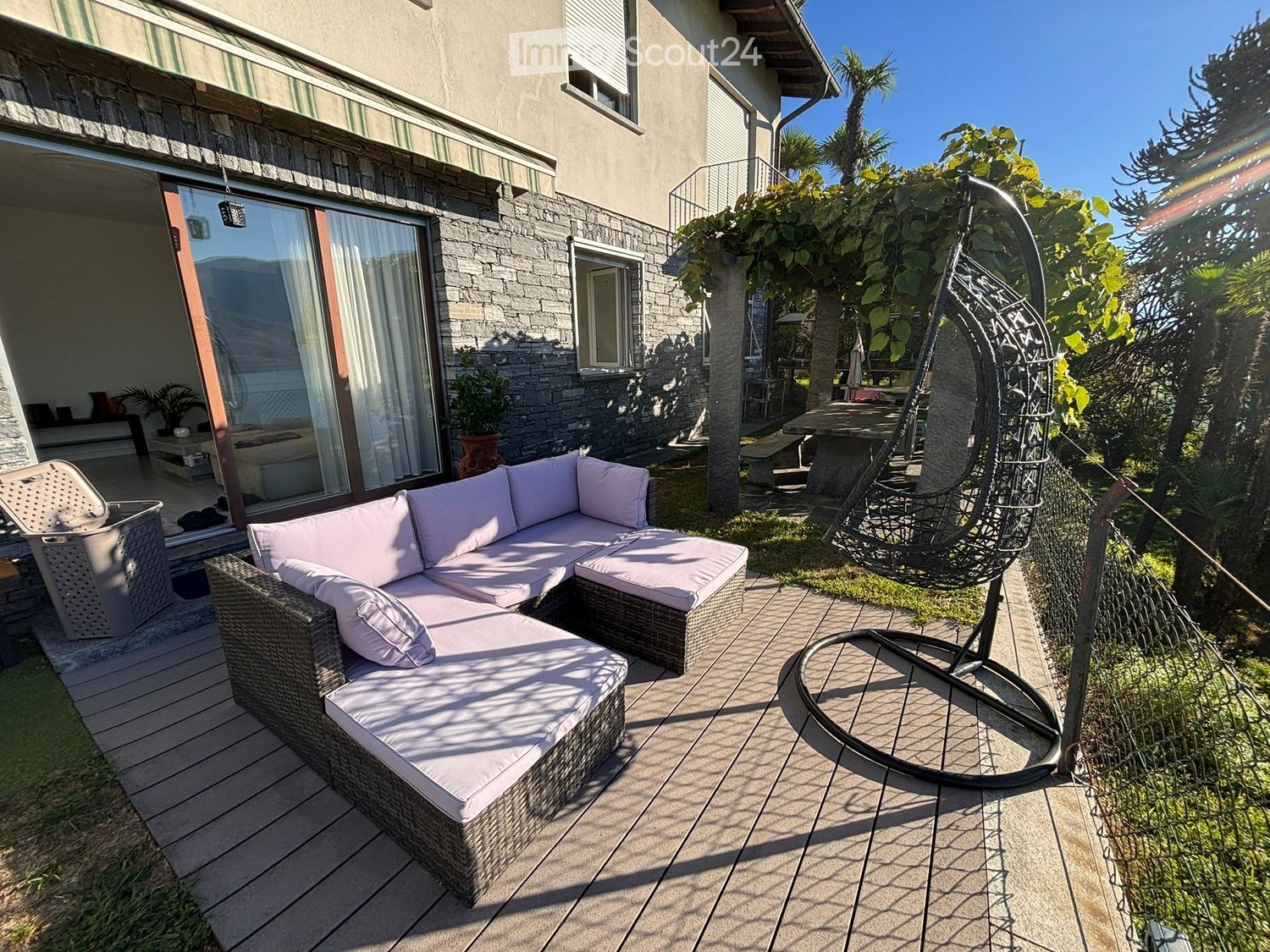 Outdoor patio with a wicker sofa, hanging egg chair, and a table surrounded by lush greenery and a modern house in the background.