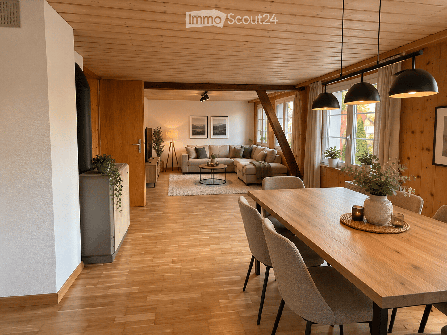 Open-plan living and dining area with wooden floors, exposed wooden beams, large windows, modern lighting, and contemporary furniture including a sofa, coffee table, dining table, and chairs.