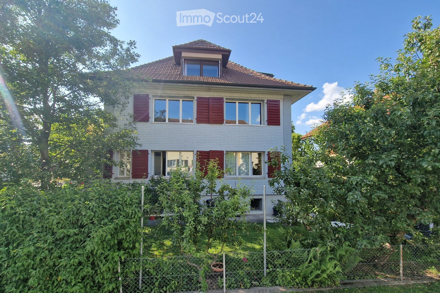 Two-story house with white brick facade, red shutters, and multiple windows, surrounded by lush greenery and a fence.