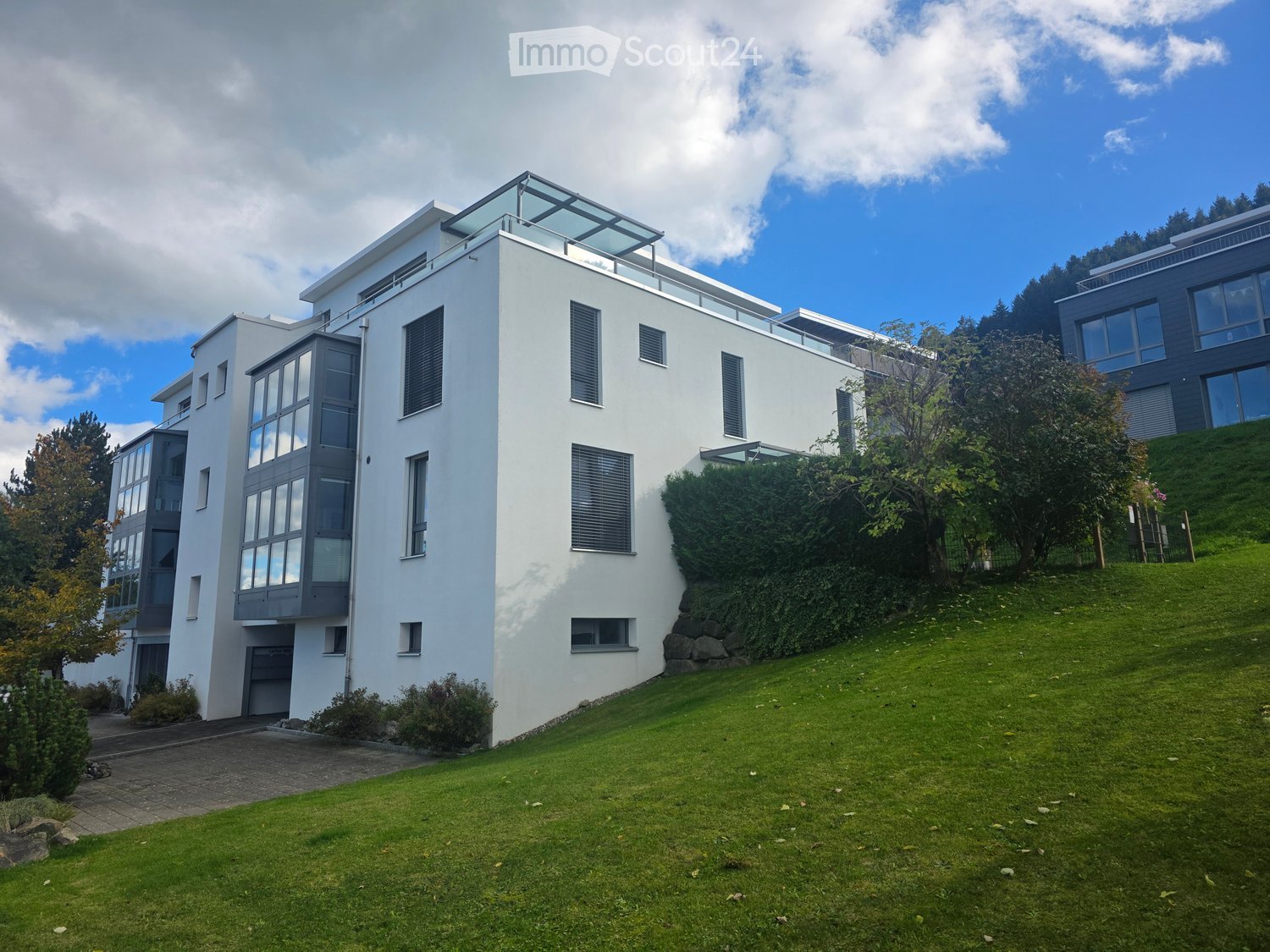 Modern two-story house with white exterior, large windows, and a flat roof terrace. The house is surrounded by a well-maintained lawn and trees.