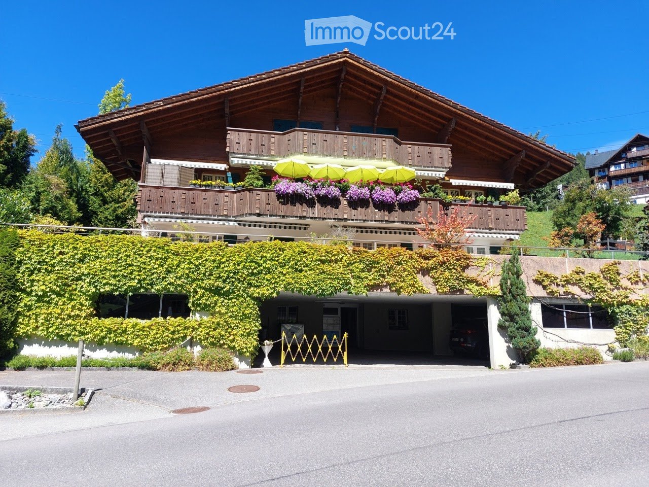A large, two-story wooden house with a steeply pitched roof, balconies adorned with colorful flower boxes, and a covered parking area partially covered by climbing plants.