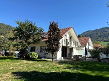 Two-story house with a red-tiled roof, white exterior, and a spacious lawn with trees and outdoor seating.