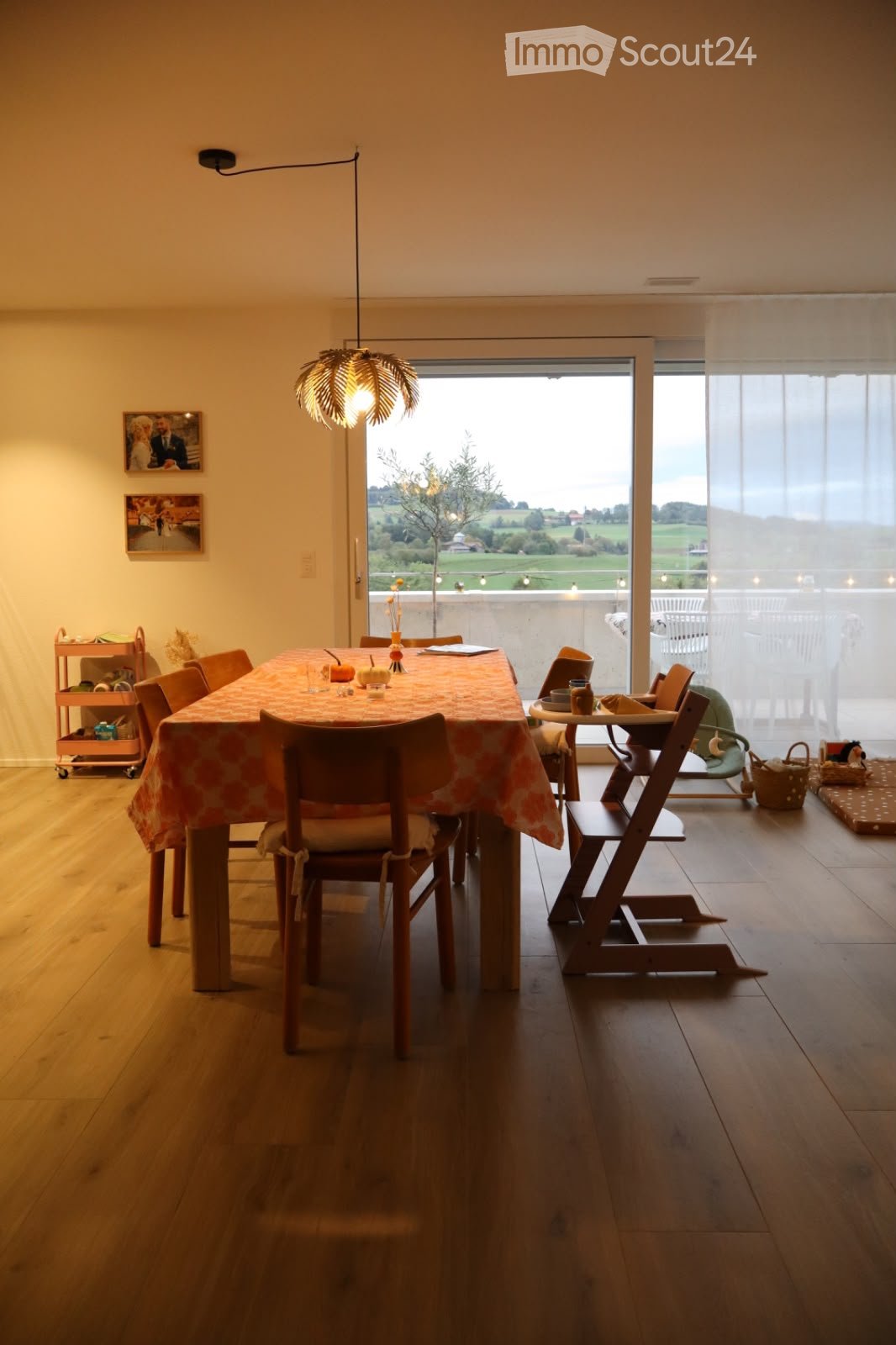 Bright dining room with wooden floors, large window offering a scenic view, dining table with chairs, baby chair, small table, and a rolling cart.