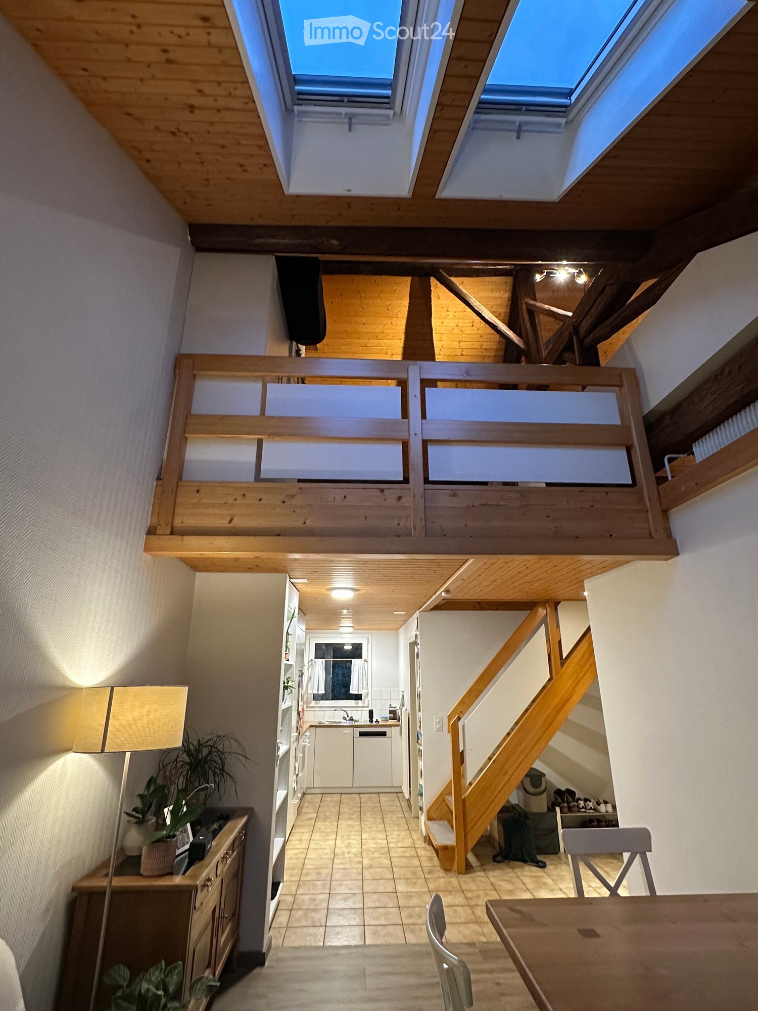 The image shows a modern, well-lit interior space with wooden beams and skylights. The main area includes a dining table with chairs, a lamp, and a cabinet with a potted plant. A staircase leads to an upper level, and a kitchen area is visible with white