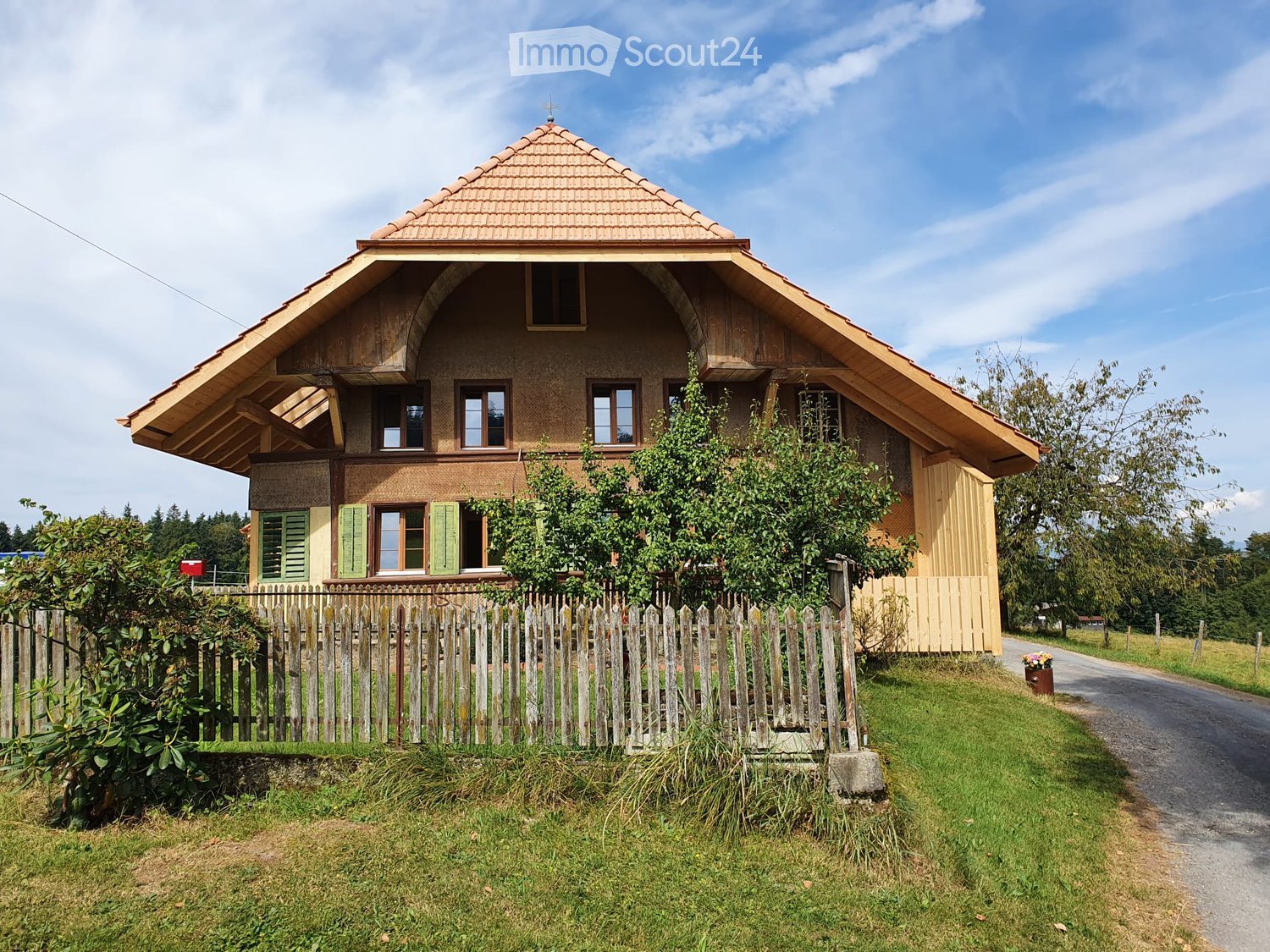 Two-story wooden house with a tiled roof, green shutters, and a wooden fence surrounding the property.