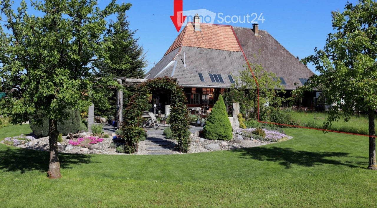 A well-maintained garden with a large house in the background, featuring a variety of plants, trees, a pergola, and a seating area.
