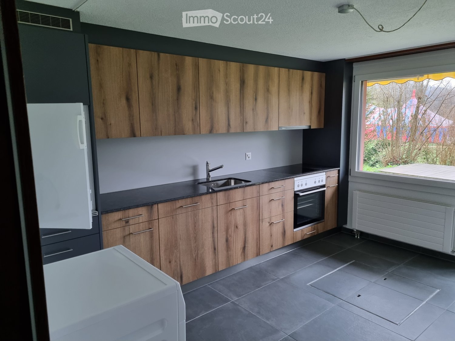 Modern kitchen with wooden cabinets, stainless steel sink, oven, and refrigerator. Grey countertops and dark tiled flooring.
