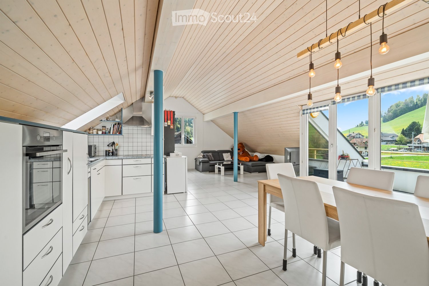 Open-plan living and dining area with white tiled flooring, wooden ceiling beams, and large windows overlooking a green landscape. Features a sofa, dining table with chairs, kitchen appliances, and pendant lights.