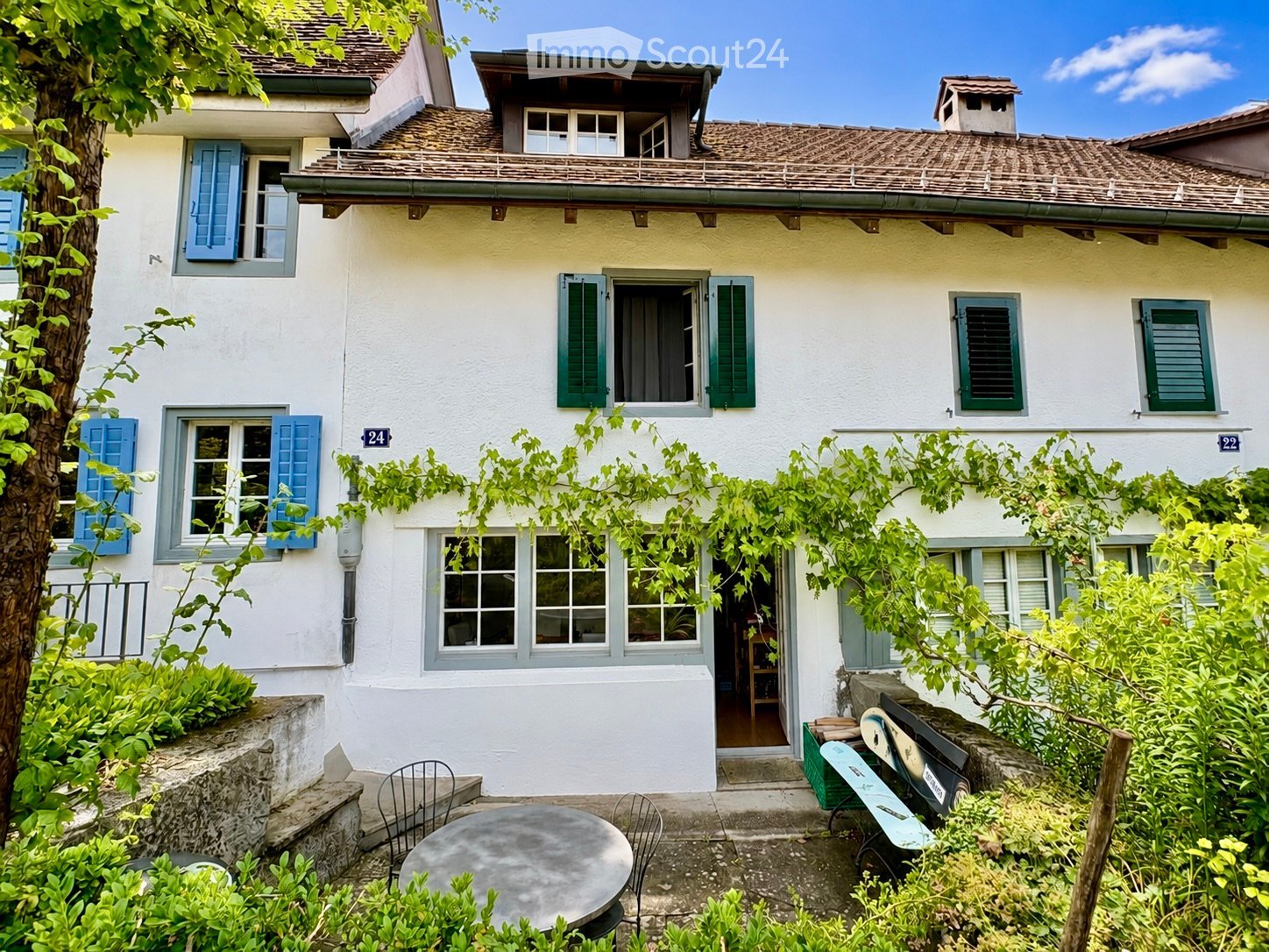 Two-story house with white walls, blue shutters, and a tiled roof. The house has multiple windows and a small balcony on the upper floor. The entrance is surrounded by greenery, with outdoor seating including a round table and chairs.