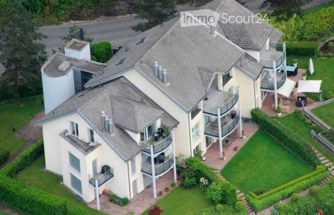 A large, modern multi-story house with multiple balconies, a well-maintained garden, and a patio area with outdoor furniture.