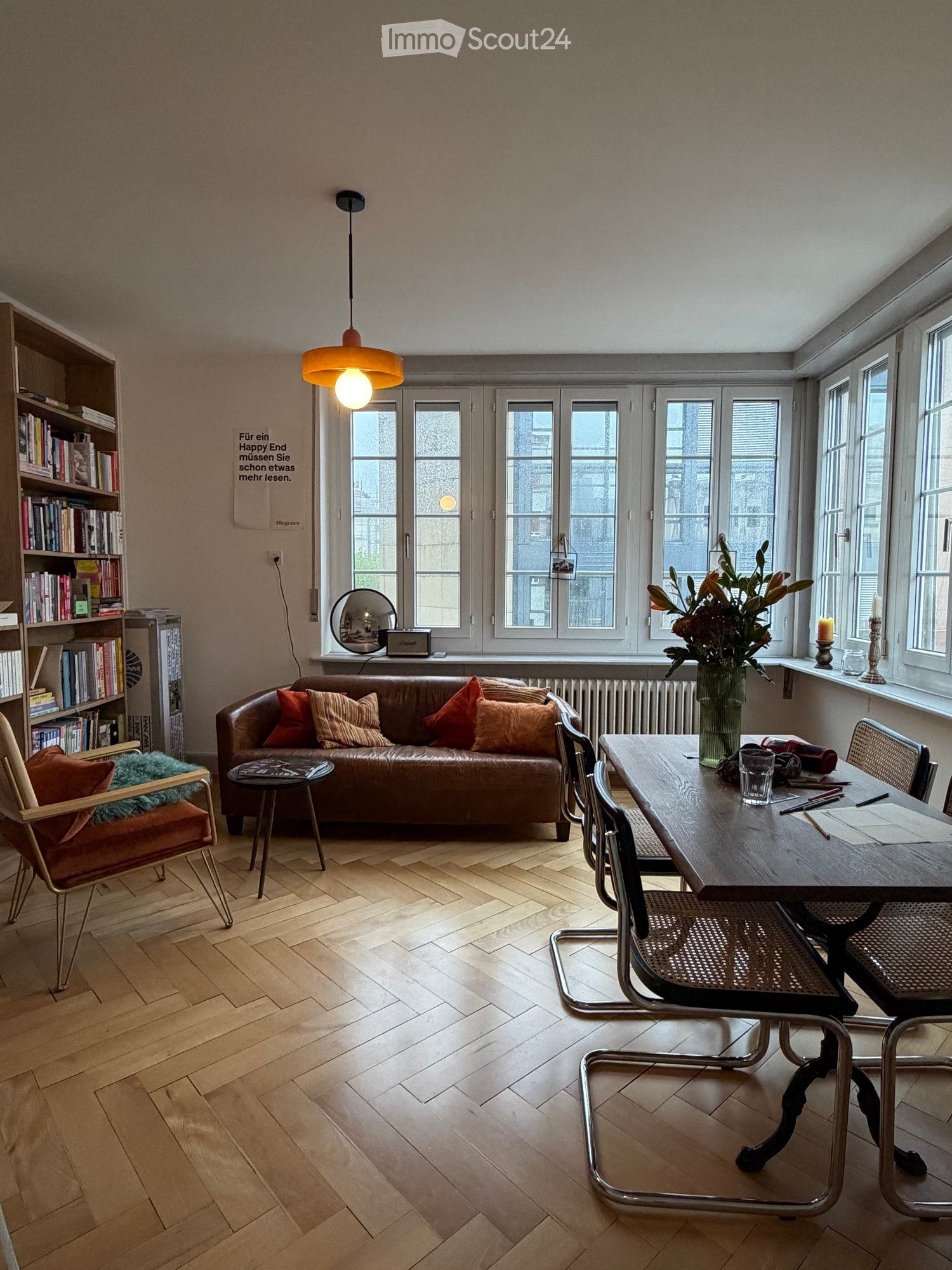 A modern living room with wooden flooring, large windows, a brown leather sofa with colorful pillows, a wooden dining table with chairs, a bookshelf, and a pendant light.