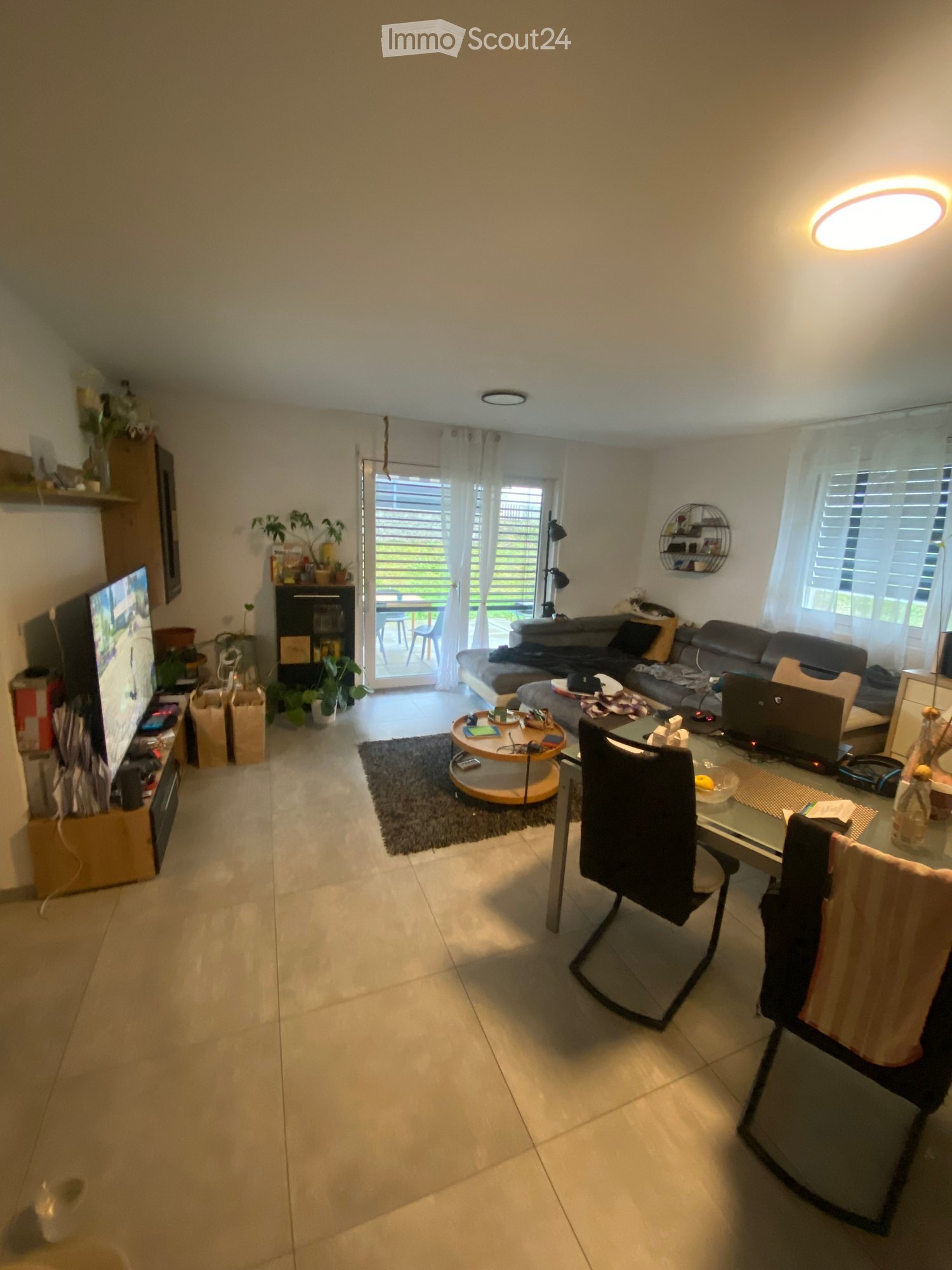 A modern living room with a sofa, coffee table, dining table, chairs, laptop, TV screen, shelves, and plants. The room features large windows with curtains and a sliding glass door leading to a balcony.