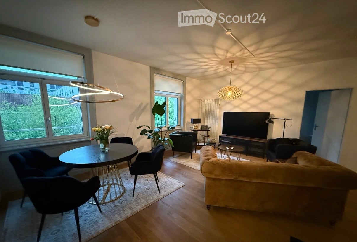 Modern living room with a cream-colored sofa, coffee table, dining table with chairs, and a television. The room features large windows with a view of greenery, pendant lights, and a small side table with a lamp.