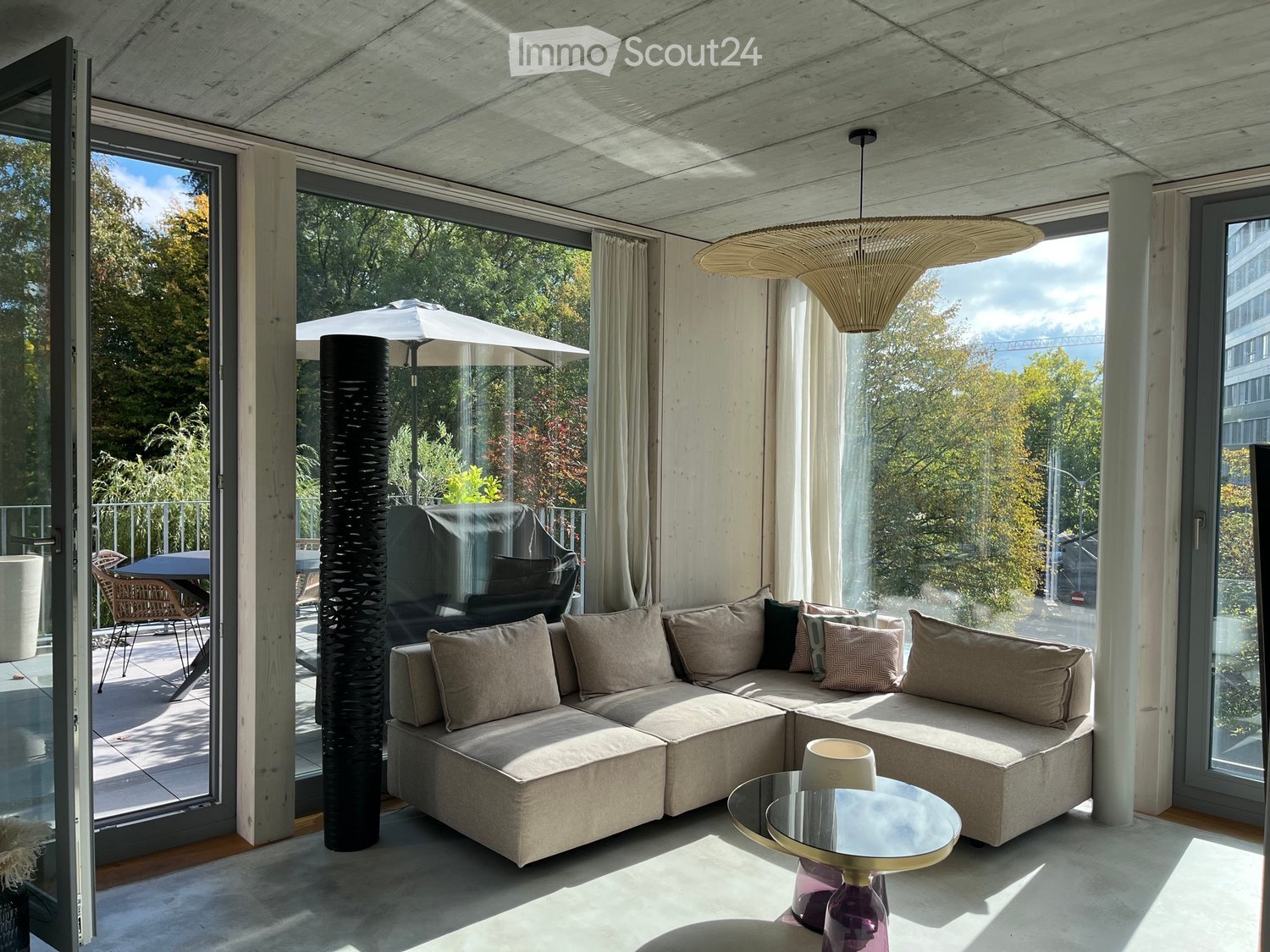 Modern living room with large windows, a sectional sofa, coffee table, floor lamp, and outdoor furniture visible through the glass doors.