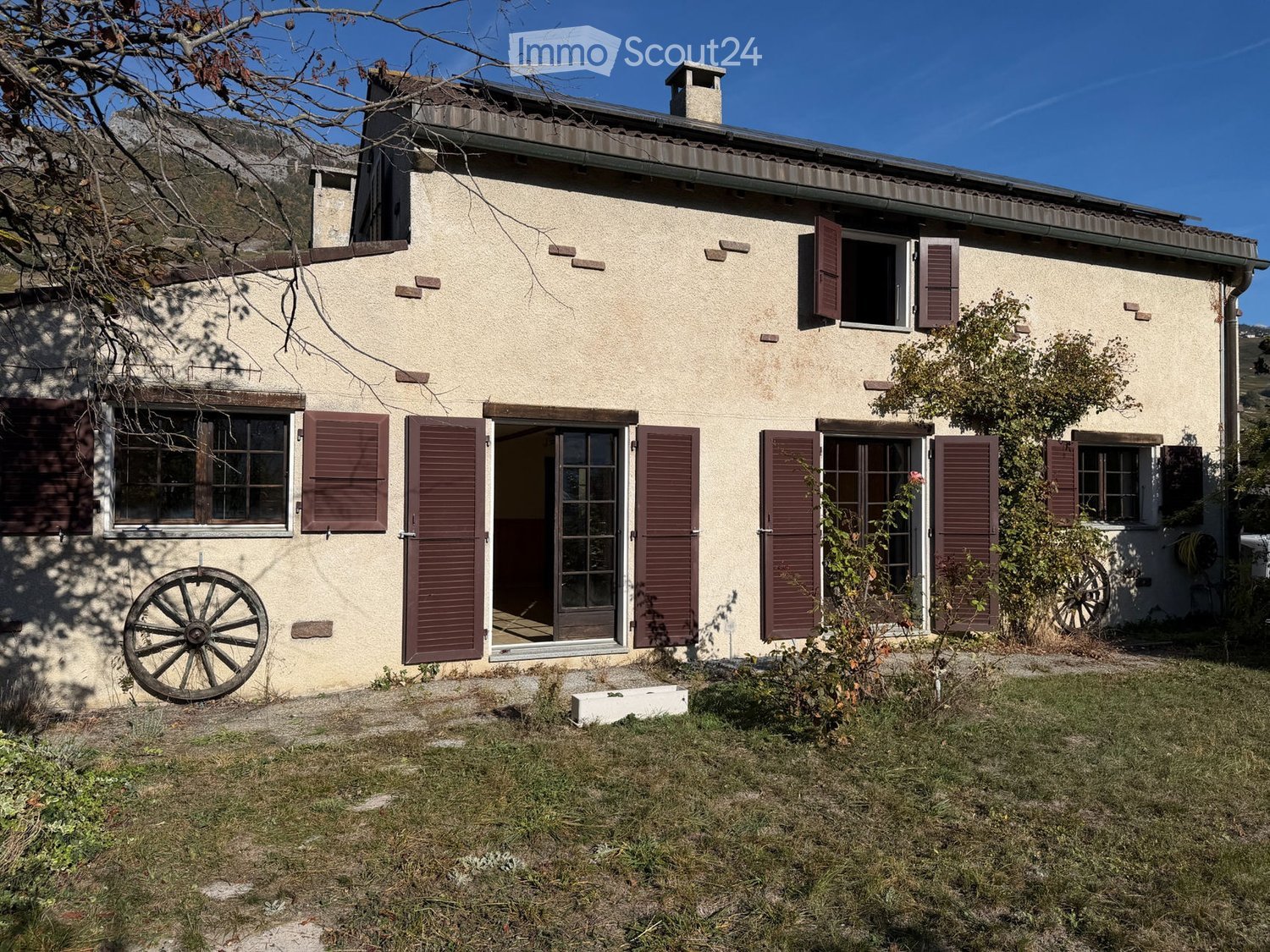 Two-story house with beige walls, red shutters, and a tiled roof. Features include a front door with glass panels, multiple windows, and a small garden area with grass and a few plants.