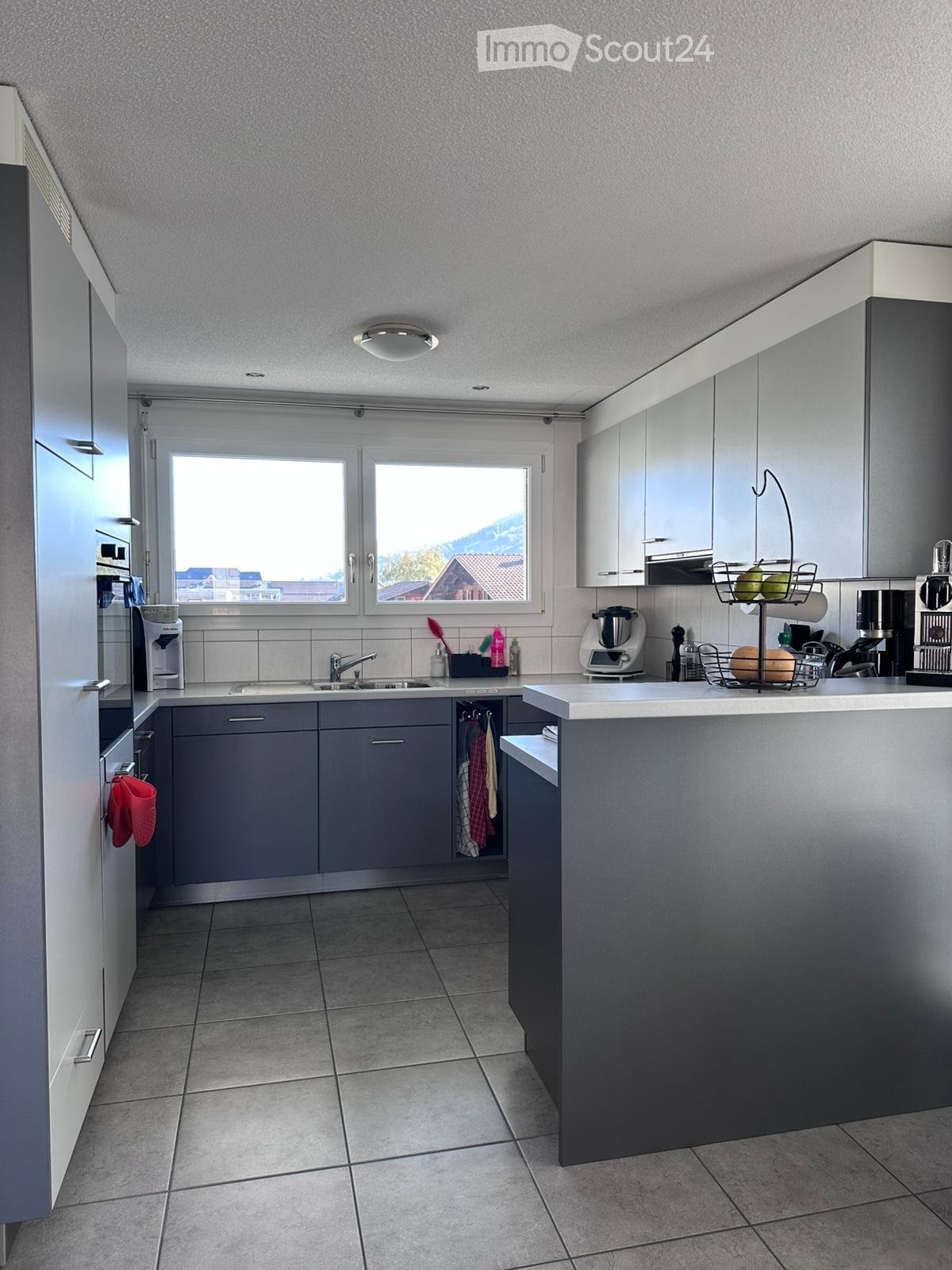 Modern kitchen with grey cabinets, white countertops, and a large window offering a scenic view. Includes a sink, coffee maker, blender, and other appliances.