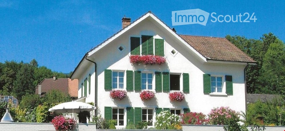 Two-story white house with green shutters, multiple windows, and flower boxes on the facade. The house has a brown tiled roof and is surrounded by greenery and a small garden area with an umbrella.