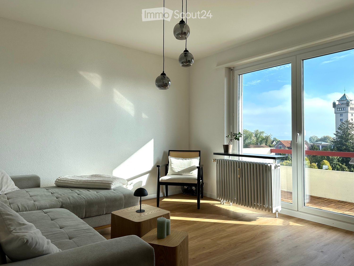 Modern living room with grey sectional sofa, wooden coffee table, armchair, radiator, and large windows offering a scenic view.