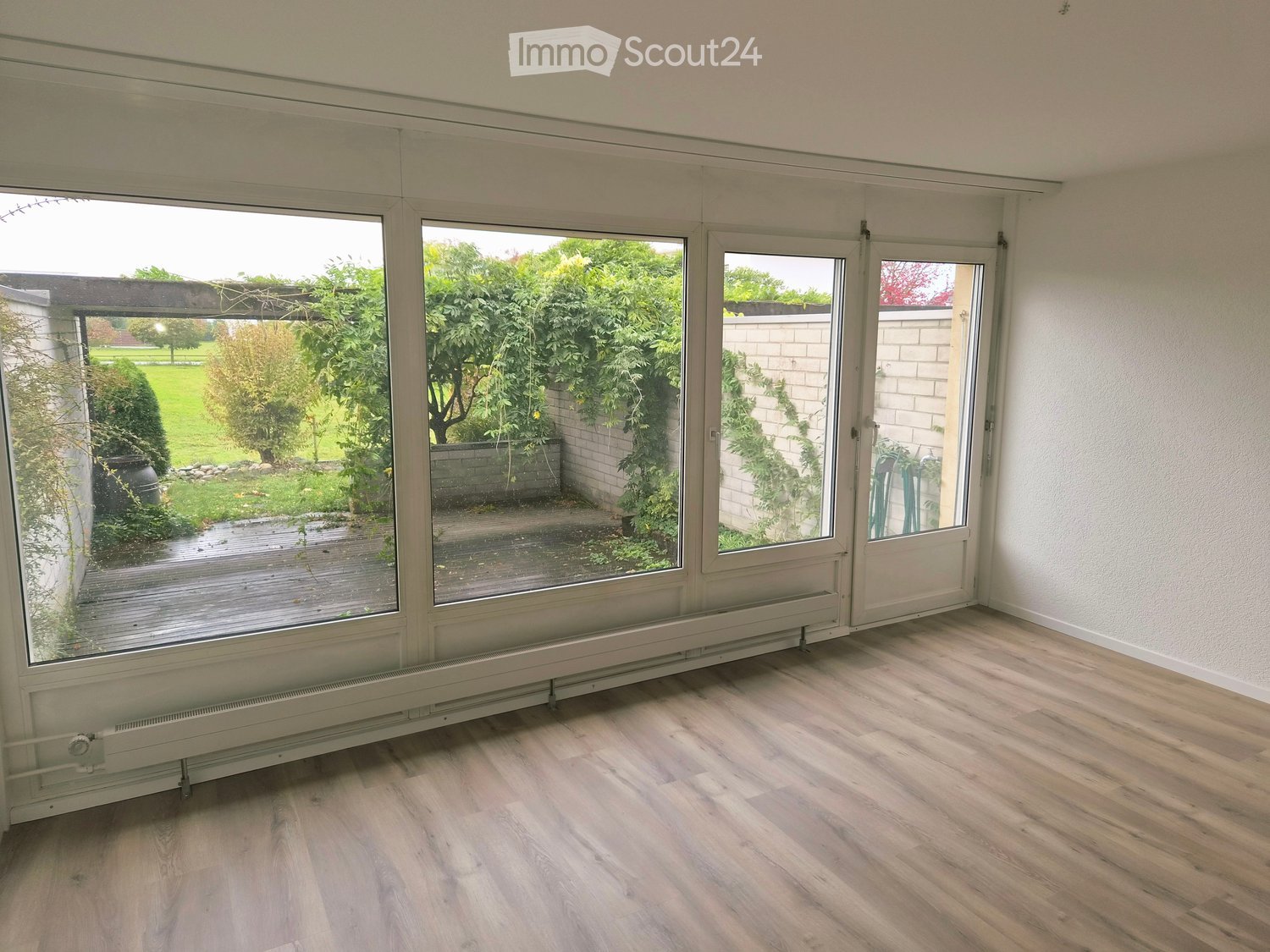 Empty room with large windows and wooden flooring, leading to an outdoor area with greenery and a paved patio.