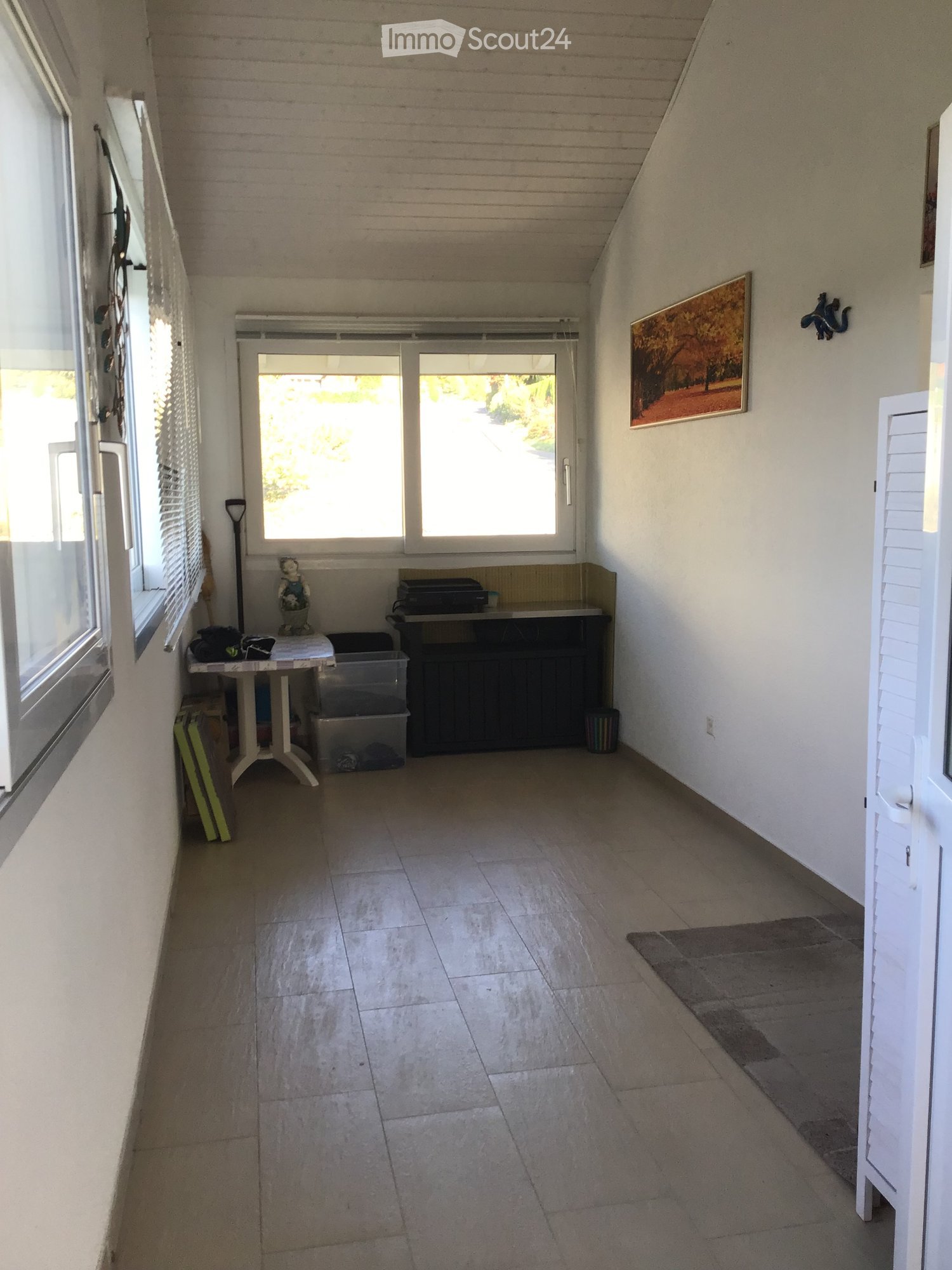 A small, well-lit room with tiled flooring, white walls, and large windows. Contains a small table with items, a cabinet, a stool, and a picture frame on the wall.
