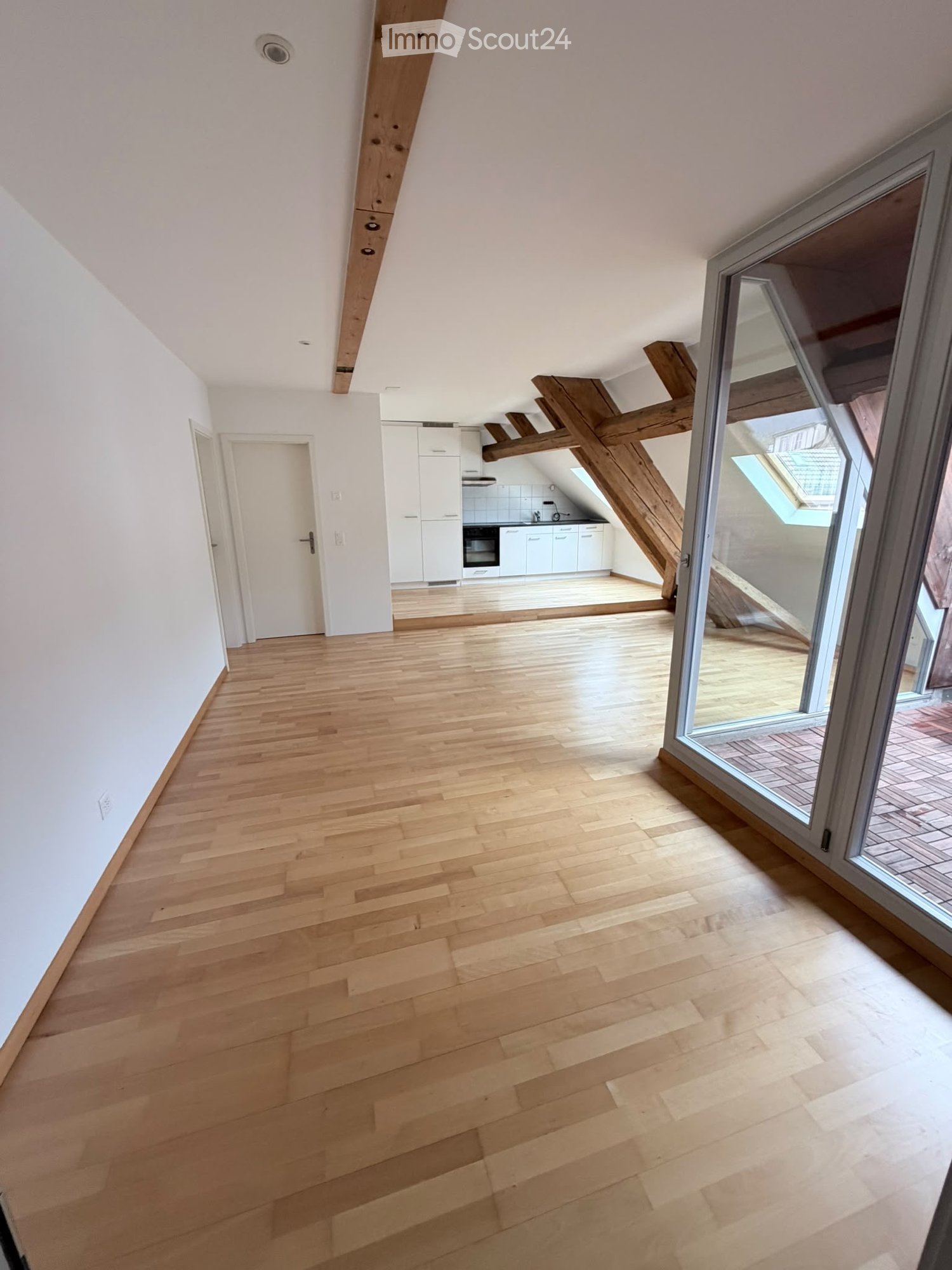 Empty room with wooden flooring, white walls, and exposed wooden beams. Features a large glass door leading to a patio and a doorway to a small kitchen area.