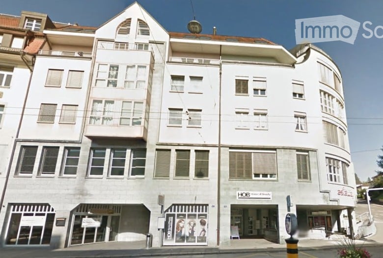Multi-story building with white facade, multiple windows, and balconies. Ground floor has commercial storefronts with signage.
