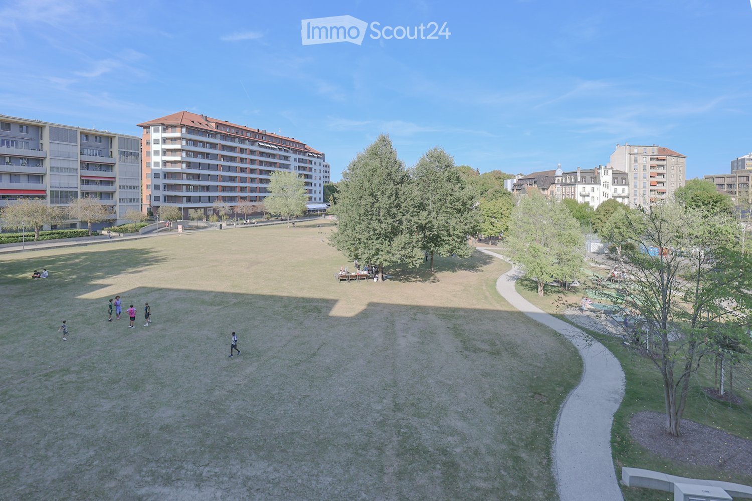 A spacious park area with grassy fields, trees, pathways, and surrounding residential buildings.