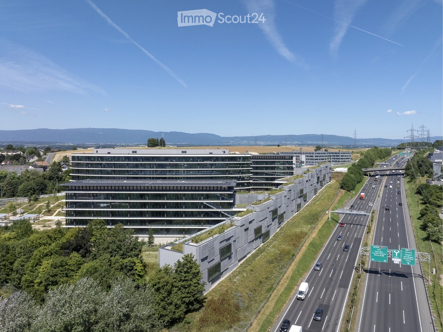 Aerial view of a modern office building complex with multiple structures, surrounded by greenery and a highway with traffic passing by.