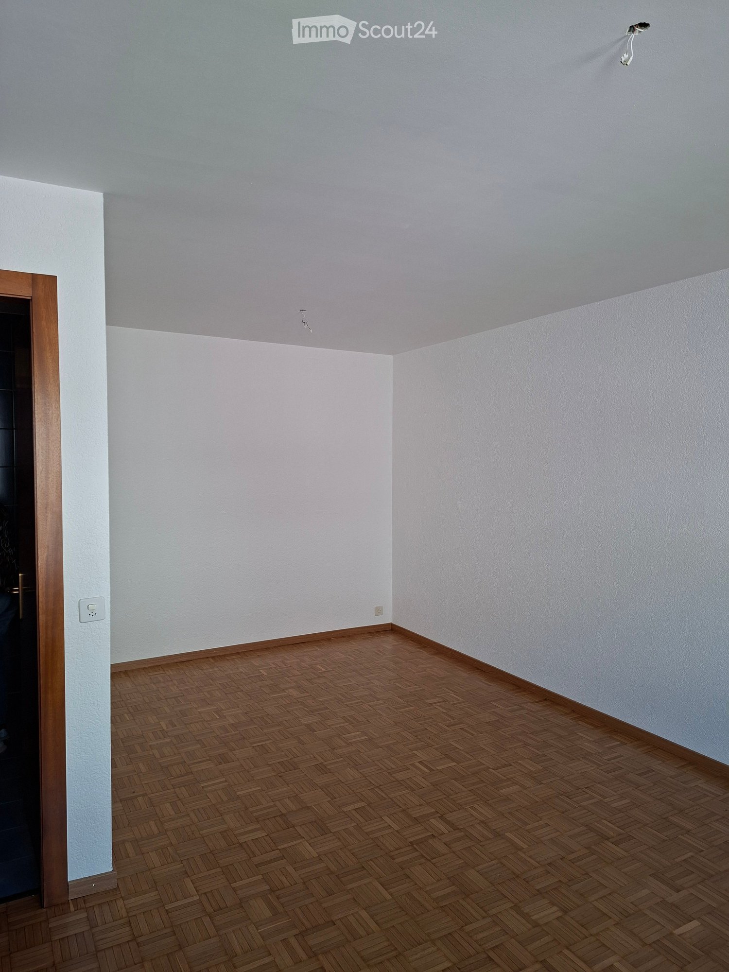 Empty room with light grey walls and a herringbone-patterned wooden floor. The room features a wooden door frame on the left and a visible electrical outlet on the wall.