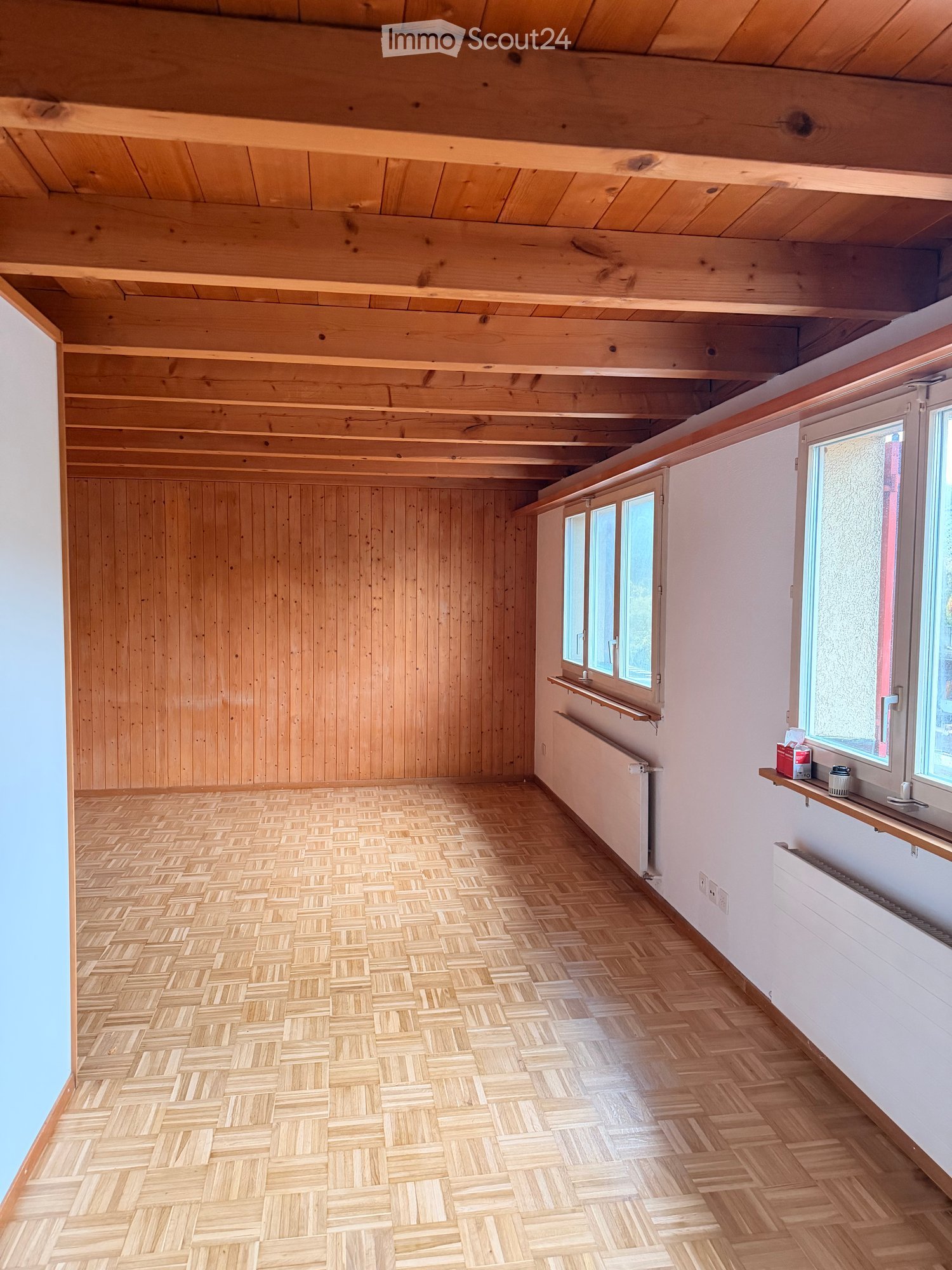 Empty room with wooden walls, herringbone wooden flooring, and exposed wooden ceiling beams. Windows with white frames and radiators are visible.