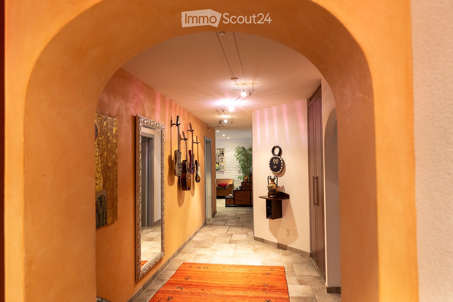A well-lit hallway with arched ceilings, warm-toned walls, and tiled flooring. Features include a large mirror, wall-mounted guitars, a small shelf with decorative items, and a glimpse of a room with a couch and plant in the background.