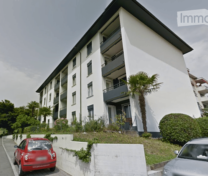 Modern multi-story apartment building with white facade, multiple balconies, and a raised ground floor. A palm tree and small garden area are visible in front of the building.
