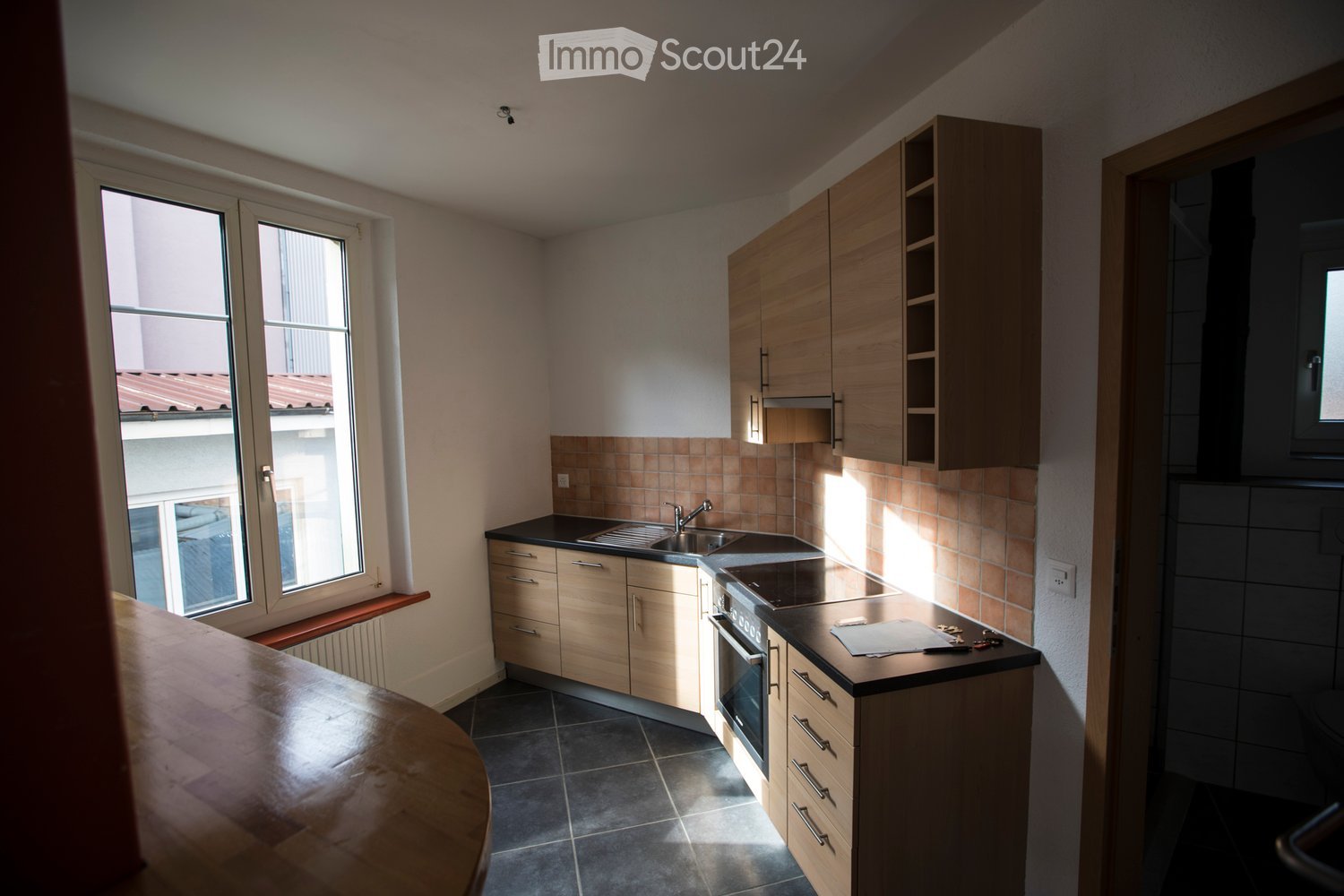 Modern kitchen with light wooden cabinets, black countertop, sink, and oven. Features large windows with a view of the roof and a wooden bar counter in the foreground.