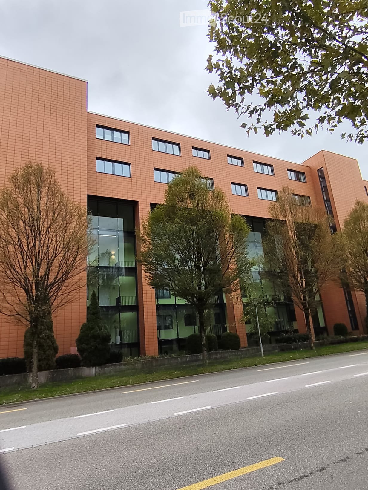 Modern two-story building with orange brick facade and large glass windows, surrounded by trees and shrubs.