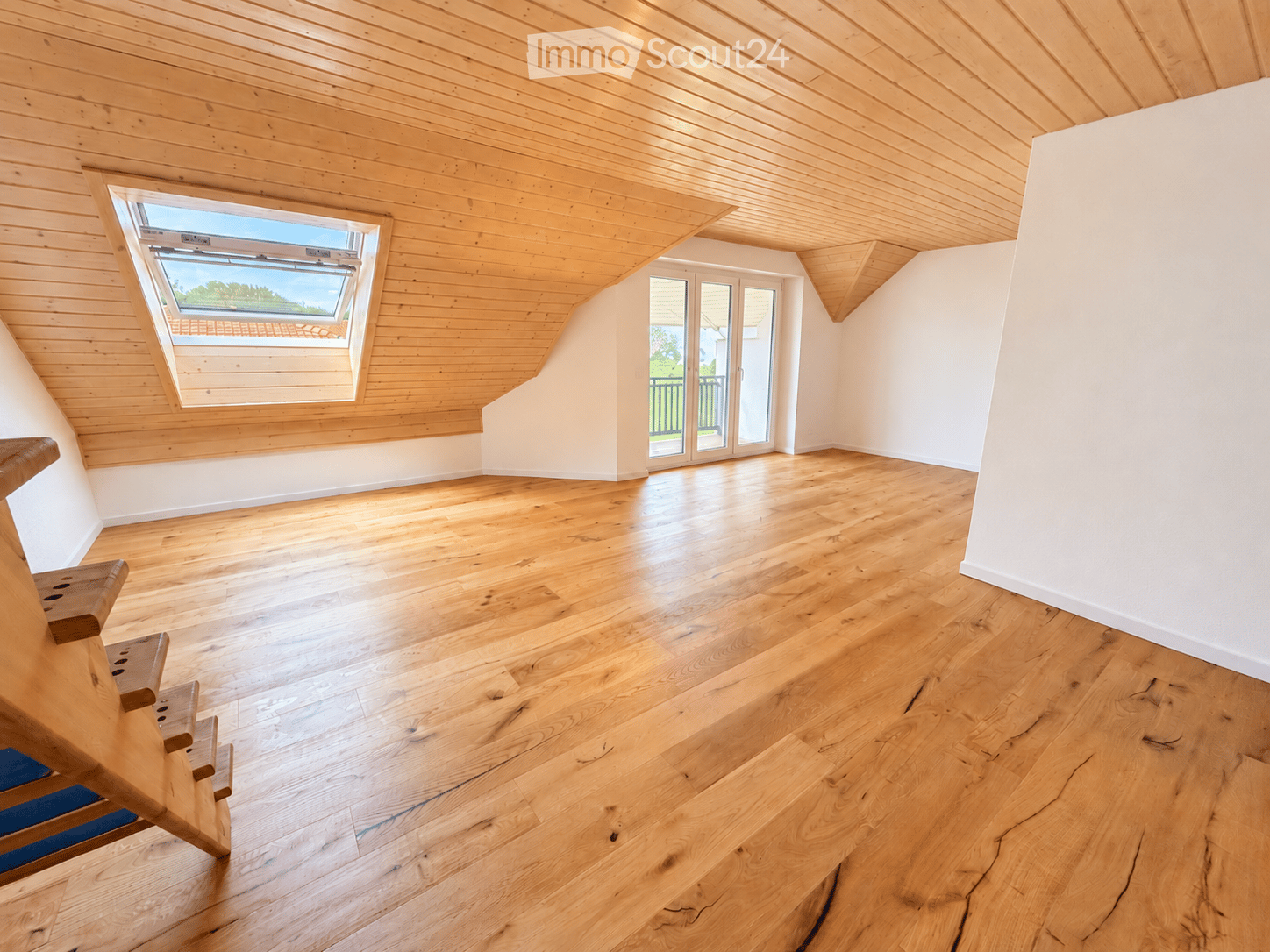 Empty attic room with wooden floors, white walls, skylight window, and sliding glass door leading to a balcony.