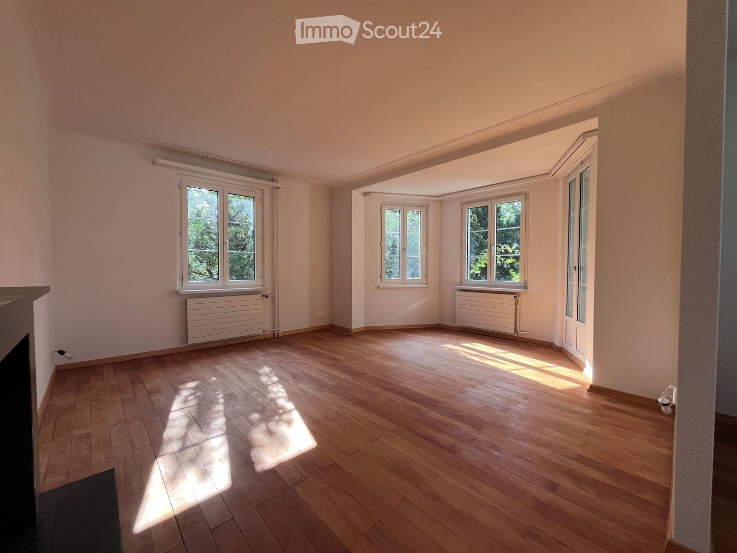 Empty room with wooden flooring, large windows, radiators, and natural light streaming in.