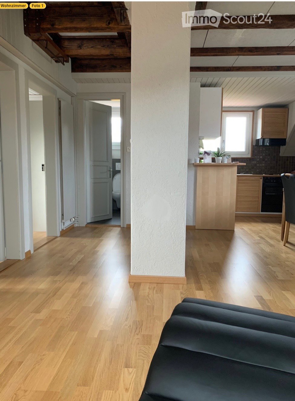 A modern, open-plan living and dining area with light wooden flooring, exposed wooden beams, a black leather sofa, a kitchen island, and a dining chair. The kitchen features wooden cabinets, a microwave, and a small plant on the island.