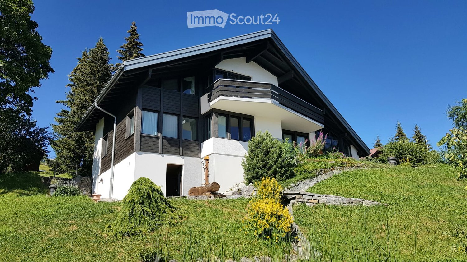 Two-story house with white walls, dark wooden accents, large windows, and a balcony on the upper level. The house is surrounded by a well-maintained lawn, shrubs, and trees.