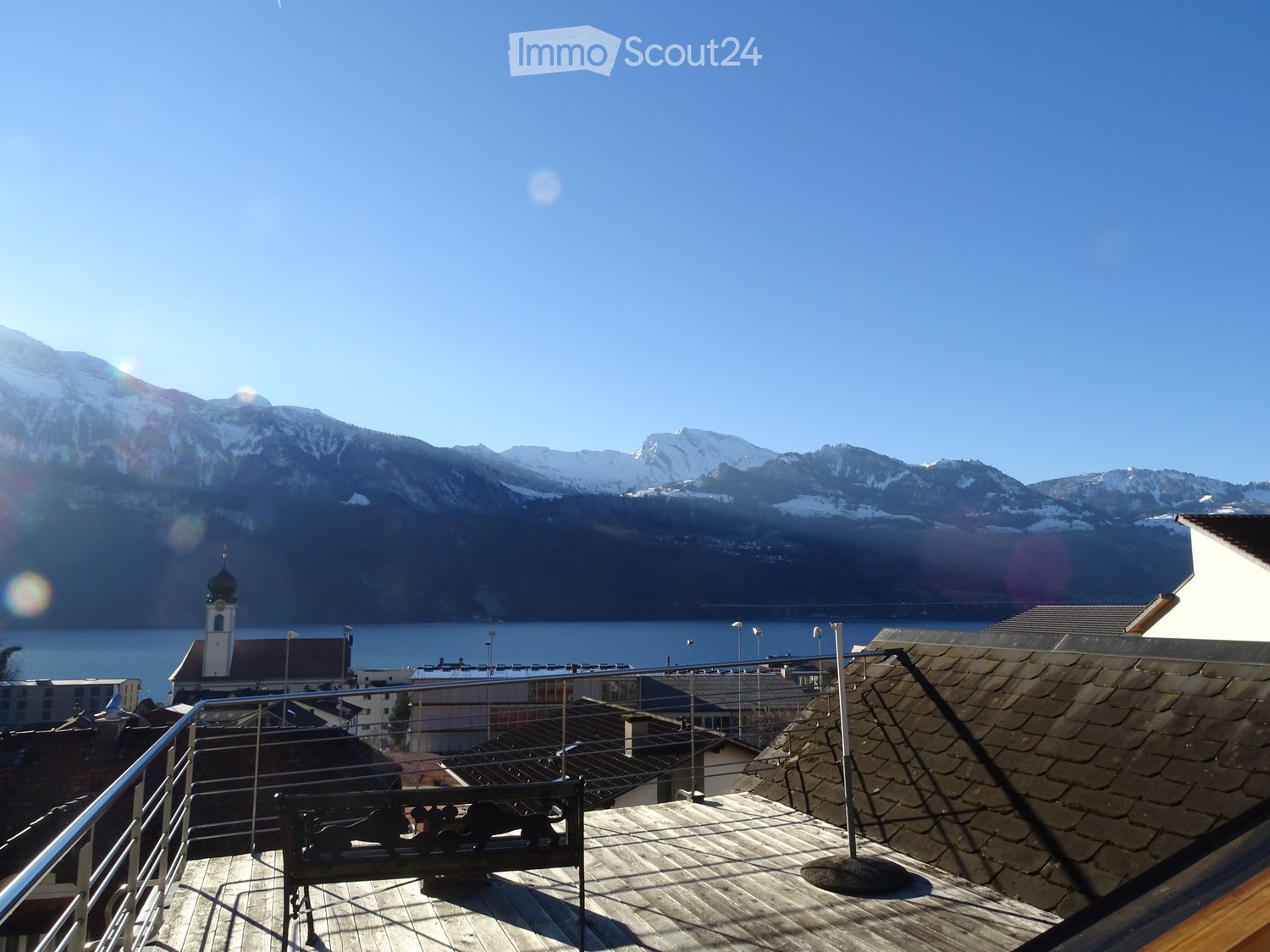 Roof terrace with wooden decking, metal railing, and a bench, overlooking a lake and snow-capped mountains.