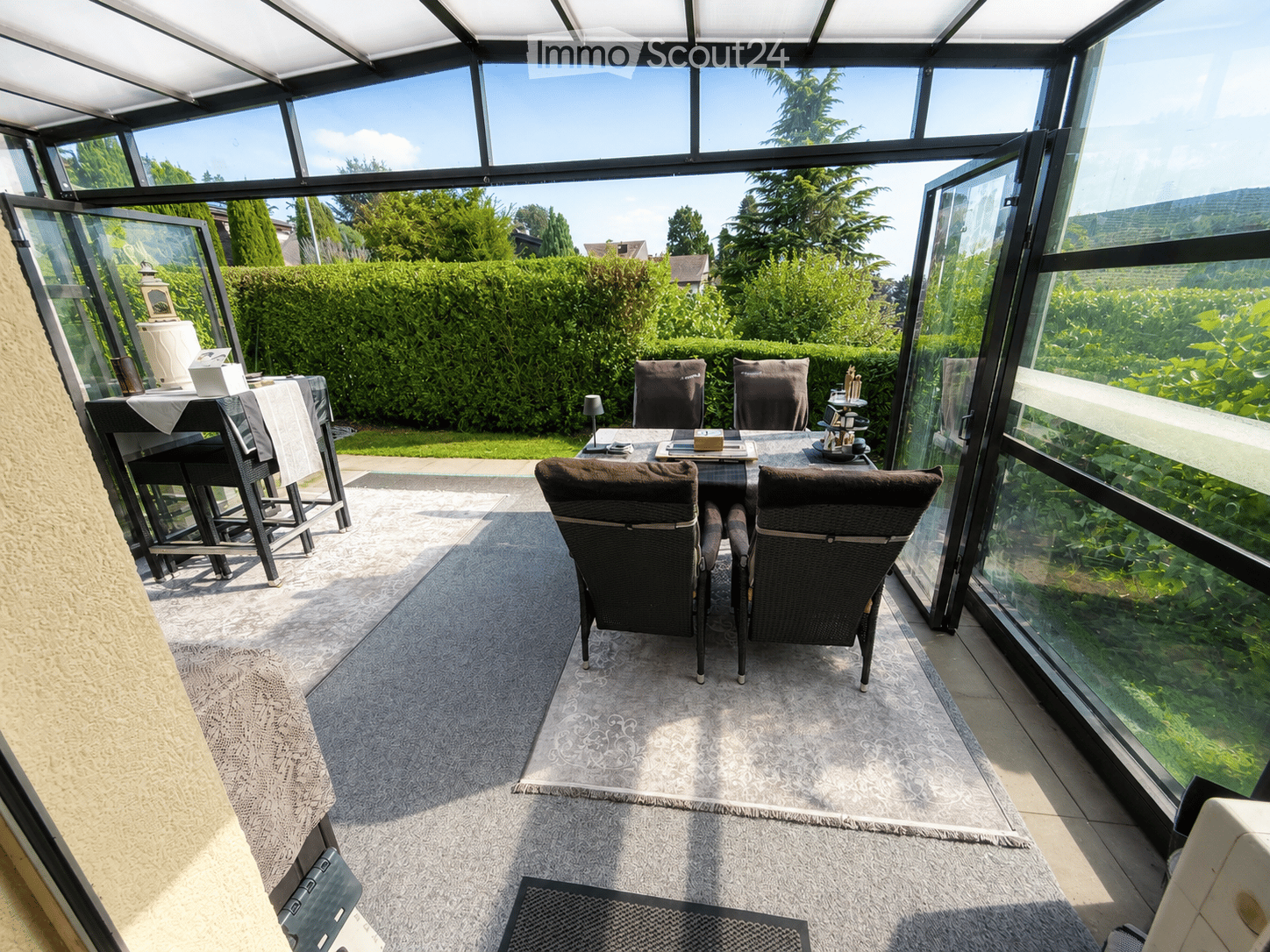 A spacious, enclosed patio with large glass walls and a retractable roof, featuring a dining table with chairs, a lamp, and a table runner, surrounded by lush greenery.