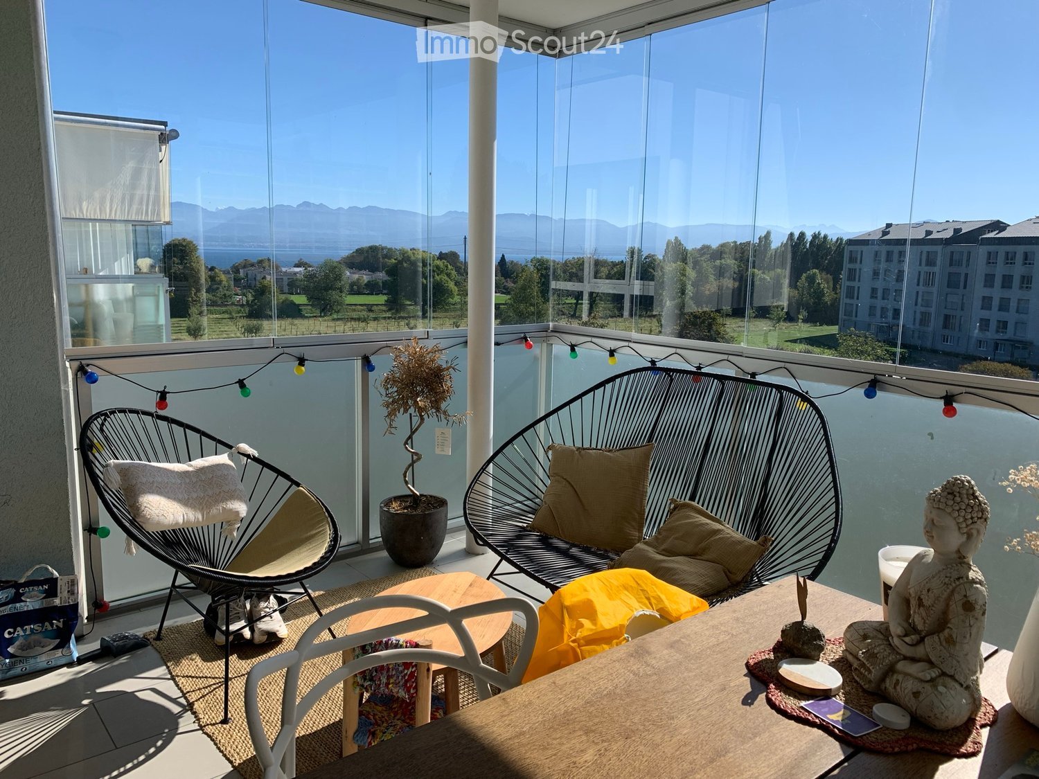Balcony with a view of mountains and water, featuring a wicker chair, a sofa, a small table, a potted plant, and a decorative statue.