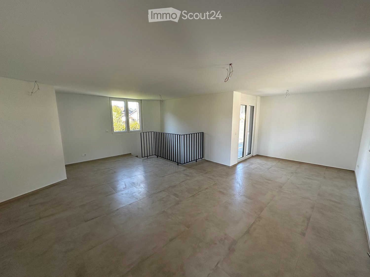 Empty room with wooden and tiled flooring, white walls, and large windows. A small black barrier is placed near the center of the room.