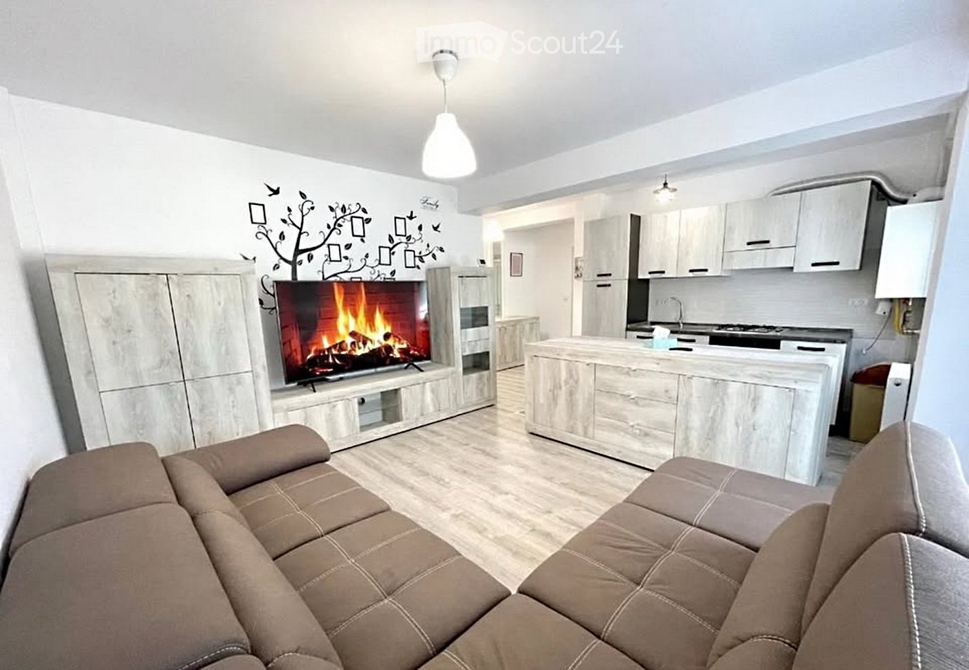 Modern living room with a beige sectional sofa, a fireplace with a TV screen, a kitchen area with white cabinets and a central island, and pendant lighting.