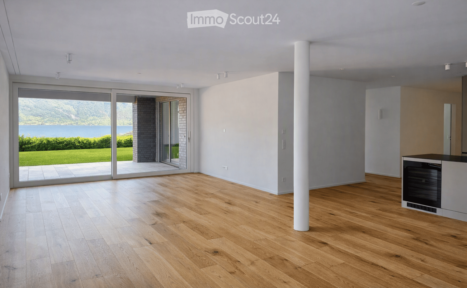 Bright, open-plan room with light wooden flooring, white walls, and large sliding glass doors leading to a view of a lake and greenery. Minimalist design with a central white column and recessed lighting.