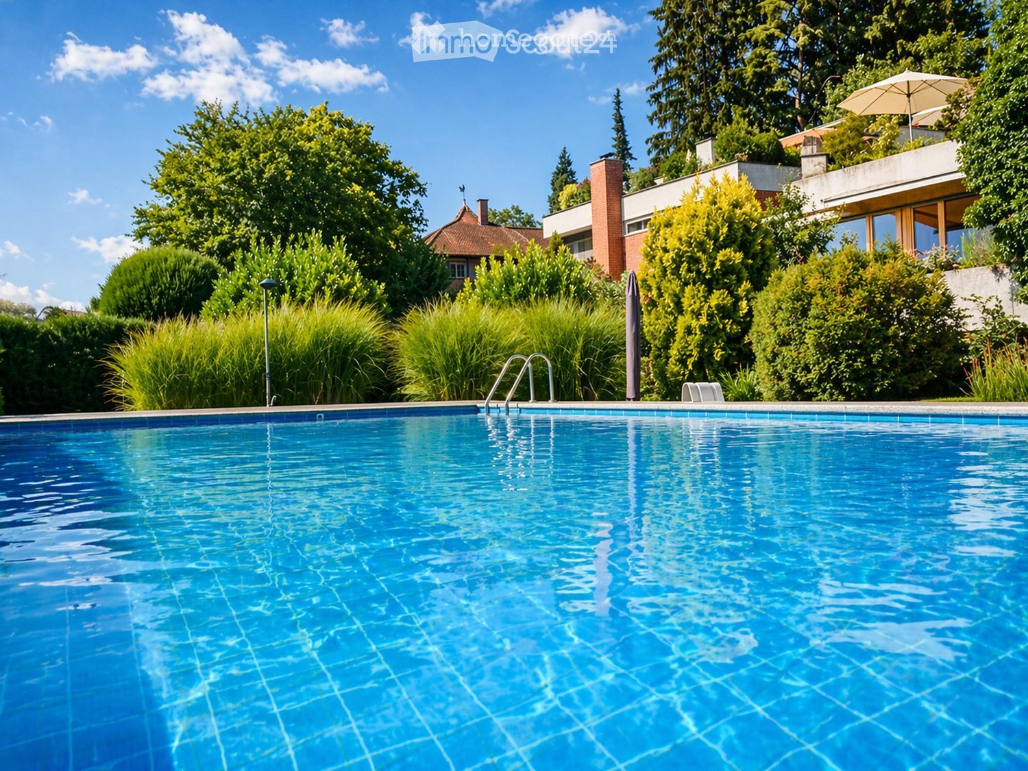 A swimming pool surrounded by greenery, with a modern building and umbrella in the background.