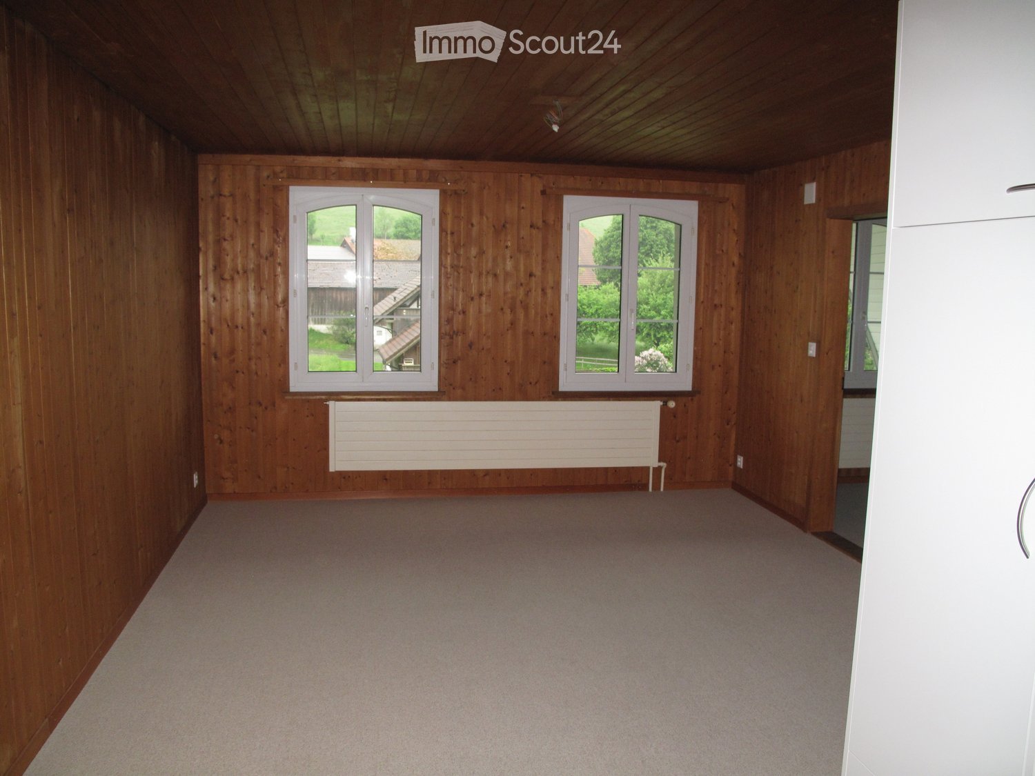 Empty room with wooden walls and ceiling, carpeted floor, two windows with white frames, and a white radiator below them.